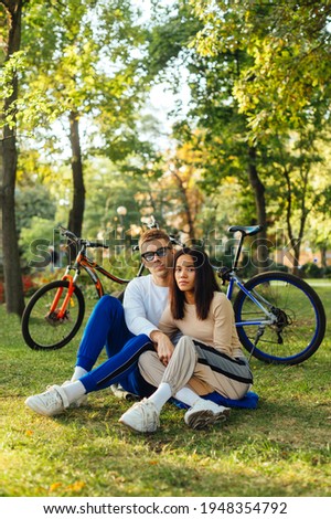 Vertical photo of a lovely beautiful couple embracing and sitting on a grass in the park. Both looking at the camera, feeling relaxed. Bicycles on the background.