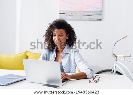 Cheerful african american teleworker using laptop near notebook and credit card