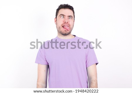 young handsome caucasian man wearing purple t-shirt against white background showing grimace face crossing eyes and showing tongue. Being funny and crazy