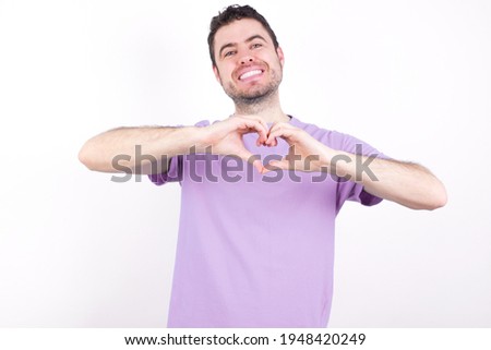 young handsome caucasian man wearing purple t-shirt against white background smiling in love doing heart symbol shape with hands. Romantic concept.