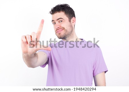 young handsome caucasian man wearing purple t-shirt against white background making fun of people with fingers on forehead doing loser gesture mocking and insulting.