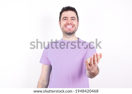 young handsome caucasian man wearing purple t-shirt against white background smiling cheerful offering palm hand giving assistance and acceptance.
