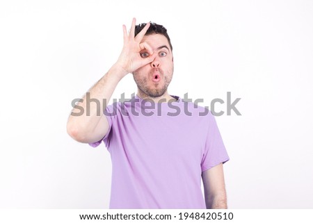 young handsome caucasian man wearing purple t-shirt against white background doing ok gesture shocked with surprised face, eye looking through fingers. Unbelieving expression.