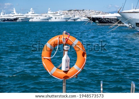 Lifebuoy concept. Orange buoy, lifebelt put on silver vertical iron. Circle lifesaver buoy support, protection, safety at Marina Flisvou, Greece. Aegean blur blue sea, ships, town background.