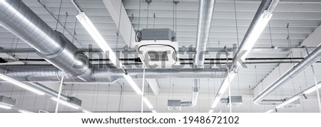 Ceiling mounted cassette type air condition units with other parts of ventilation system (tubes, cables and vents) located inside commercial hall with hanging lights and other construction parts. Royalty-Free Stock Photo #1948672102