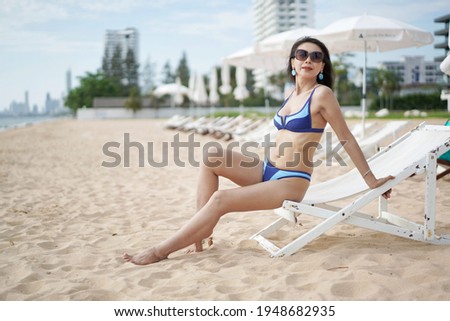 Beautiful young woman in blue bikini wearing sunglasses sitting on chair at beach, holiday activities.