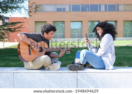 Young latin man playing the guitar and his friend recording him with the smartphone at the university campus. University life, millennial generation.