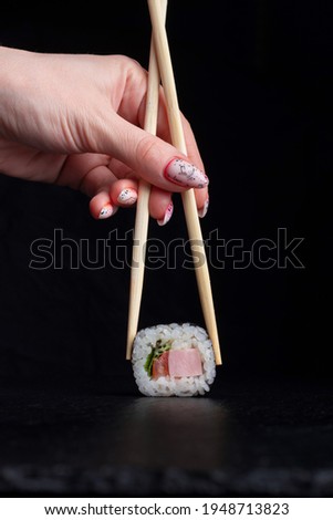 A woman's manicured hands hold wooden sticks of a classic salmon roll on a black background. Space for text, flatlay