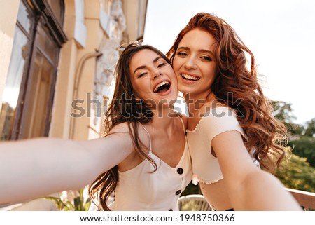 Cool ladies in white dresses take selfie on balcony. Adorable curly girls in stylish outfits laugh and take pictures together.