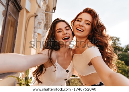 Pretty women have fun and take selfie on great terrace. Cute ladies in white summer clothes hugging and posing for camera