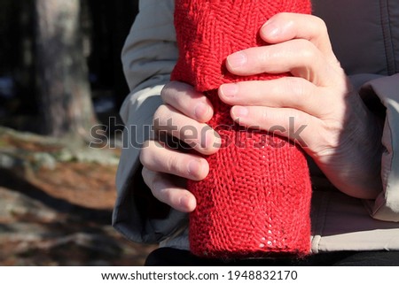 Hands of an elderly holding a thermos bottle covered by red wool sock to keep the coffee or tea warm. The photo is taken in nature. Selective focus.
