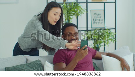 Happy mixed race couple talk laugh looking at smartphone using funny apps sit on couch. Smiling man and woman relaxing at home having fun in social media on cellphone online watching video on phone.