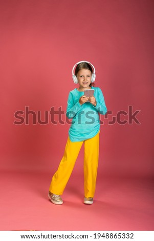 Little Beautiful Baby Girl Pink Background Bright Clothes Yellow Pants Turquoise 

Blue Shirt Wearing White Headphones Listening To Music With Smartphone In Hand