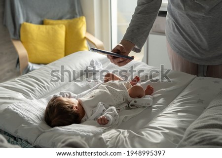 Unknown woman caucasian mother using mobile phone taking photos of her newborn baby boy or girl infant lying on the bed at home in day - modern motherhood and growing up concept