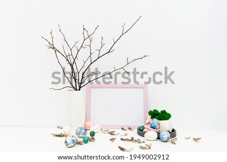 Blank photo frame for text, mockup with copy space for Easter holiday. Hand painted in trendy colors Easter eggs, geometric vase on tray with moss, feathers, branches, Easter bunny. Front view image.