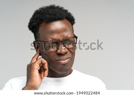 Sick African American millennial man suffering from tinnitus, throbbing earache, tired of noise. Irritated black frowning male in glasses touching painful ear, isolated on grey studio background. Royalty-Free Stock Photo #1949022484