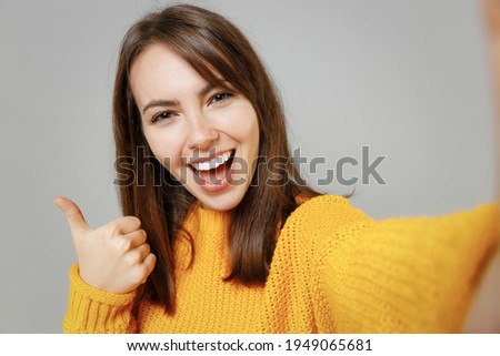 Close up young smiling happy positive attractive woman 20s in casual knitted yellow sweater do selfie shot on mobile cell phone show thumbs up like gesture isolated on grey background studio portrait.