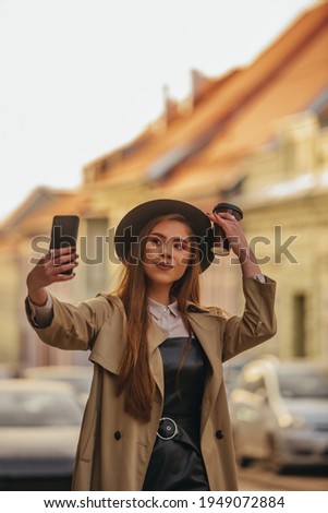 Young fashionable woman taking selfie with her smartphone while out in the city wearing coat and a hat