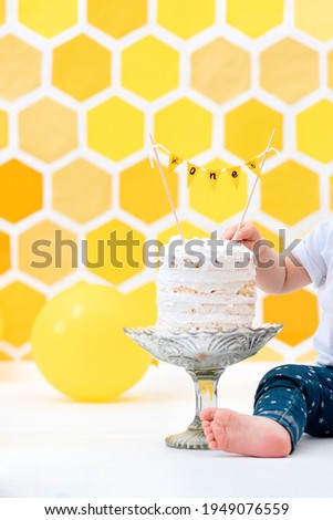 First birthday. The kid reaches for the birthday cake. Close-up. Yellow background with a decor in the form of a honeycomb and balloons. Vertical.