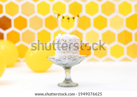 White birthday cake lying on a cake stand. Yellow background with a decor in the form of a honeycomb and balloons. Copy space.