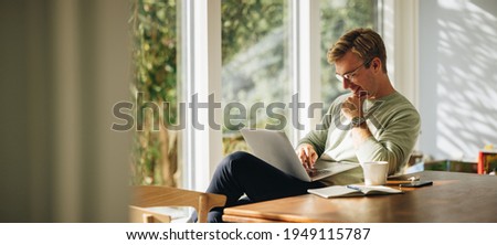 Young man using laptop and smiling at home. Man sitting by table working on laptop computer.