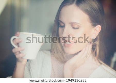 Beautiful young woman is drinking coffee. Attractive woman in a cozy cafe or at home. Pretty woman with a cup of cappuccino in her hands. Close-up portrait. Soft focus and shallow depth of field.