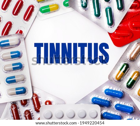The word TINNITUS is written on a light background surrounded by multi-colored packages with pills. Medical concept