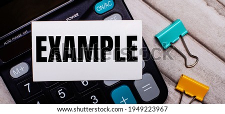 Calculator, multi-colored paper clips and a white card with the inscription EXAMPLE on a light wooden table. Stationery and calculator.