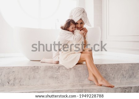 Little daughter hugs mom. Mother and daughter in white coats in a light bathroom. Hygiene, beauty and happy family concept