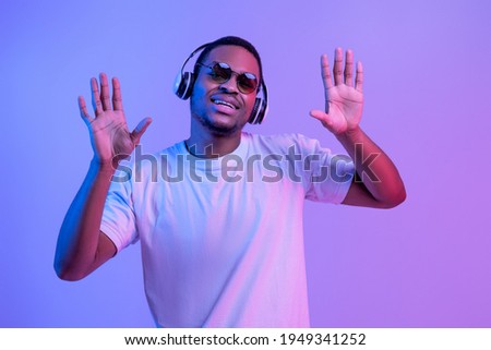 Feel The Rhytm. Cheerful Black Guy In Wireless Headphones Listening Favorite Music And Dancing, Millennial African American Man Having Fun Under Neon Light Over Purple Background, Free Space