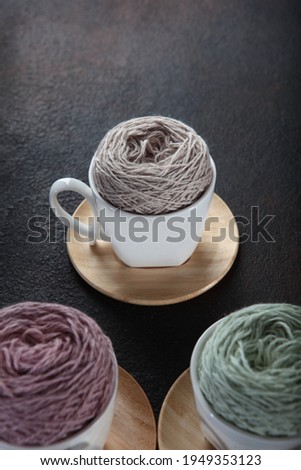 Colorful balls of yarn in white ceramic cups. White cups on wooden saucers. Hobby concept. Hand knitting.Free space. Top view. Dark background. Vertical photo.Out of focus.