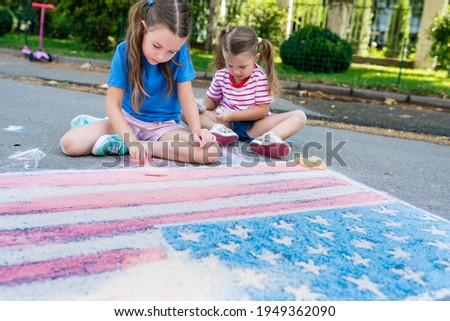 Girls drawing American flag with colored chalks on the sidewalk near the house on sunny summer day. Kids painting outside. Creative development of children.  Independence Day concept