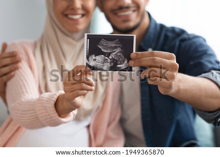 Closeup shot of happy pregnant muslim couple demonstrating baby sonography photo while relaxing together at home, showing first photo of their child, enjoying future parenthood, cropped image Royalty-Free Stock Photo #1949365870