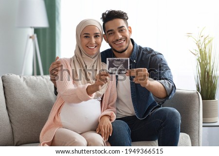 Pregnant Islamic Lady In Headscarf And Her Husband Showing Baby Sonogram Picture At Camera While Sitting On Couch At Home, Loving Muslim Family Demonstrating Ultrasound Scan And Smiling, Free Space