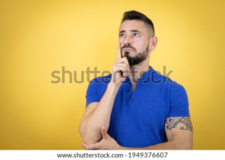 Handsome man with beard wearing blue polo shirt over yellow background serious face thinking about question with hand on chin, thoughtful about confusing idea