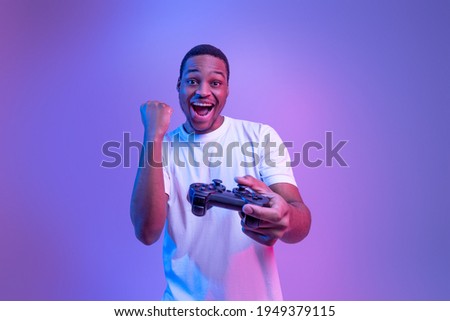 Video Gaming Concept. Euphoric African American Guy With Joystick Celebrating Game Win, Cheerful Black Man Raising Fist And Exclaiming With Excitement, Standing In Neon Light Over Purple Background Royalty-Free Stock Photo #1949379115