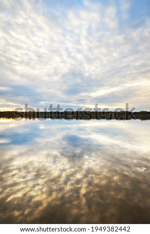 Blue sky with glowing golden sunset clouds above the forest lake. Symmetry reflections on the water, natural mirror, texture, background. Idyllic rural scene. Nature, environment, weather