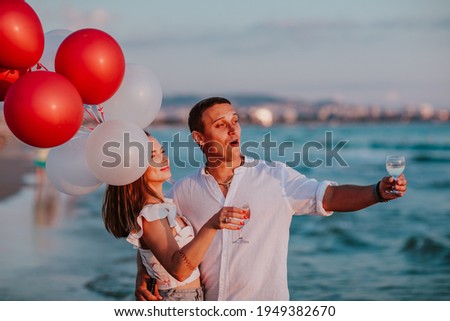 Young pretty woman and handsome man in white summer clothes hugging and celebrating together with champagne and balloons on a sea coast. Copy space.