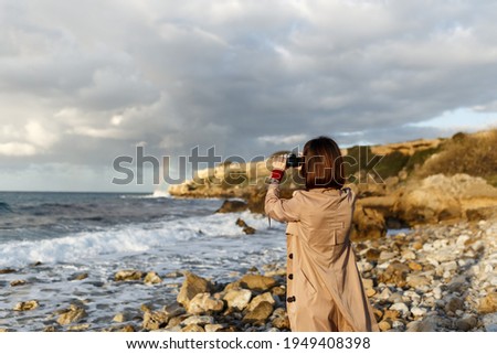 stylish hipster girl traveler on stone shore at winter wearing coat. Fashionable young woman with short red hair makes photo of ocean sea rainbow using camera.
