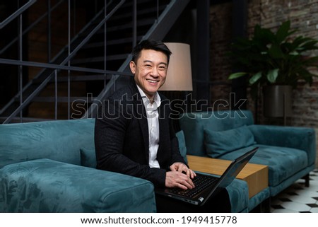 Successful Asian businessman, in a black business suit, works on a laptop, relaxes in a restaurant or hotel, and in a stylish office explains a new business strategy via video link Royalty-Free Stock Photo #1949415778
