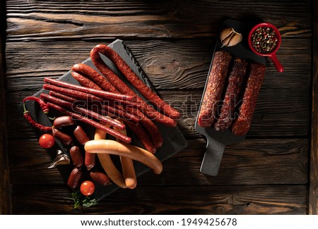 Hunting, smoked sausages of different sizes, sausages, fresh tomatoes, peppers in a saucepan and dried, garlic and parsley lie on slate boards on a wooden, brutal dark-style background. Top view..