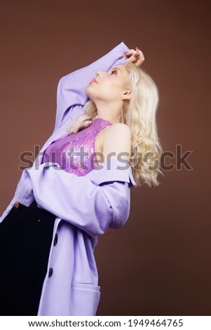 A girl in a purple jacket on a brown background in 70s, 80s, 90s style makeup, long silver earrings. The blonde is dancing, posing, fashion. A handsome woman.