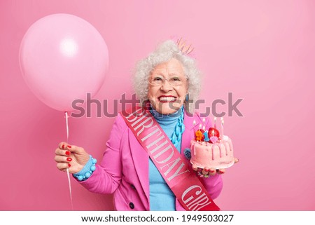 Studio shot of happy wrinkled female pensioner with bright makeup smiles toothily holds festive cake with burning candles has festive mood carries inflated balloon isolated over pink background