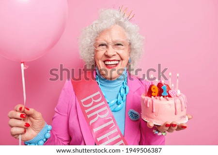 Special occasion age and festivity concept. Happy smiling wrinkled senior woman holds festive strawberry cake inflated balloon prepares for party or birthday celebration expresses good emotions Royalty-Free Stock Photo #1949506387