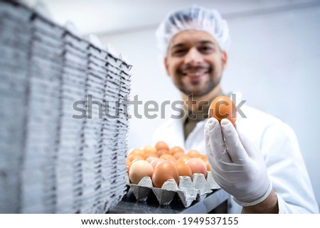 Food factory worker standing by industrial eggs packaging machine and holding an egg.