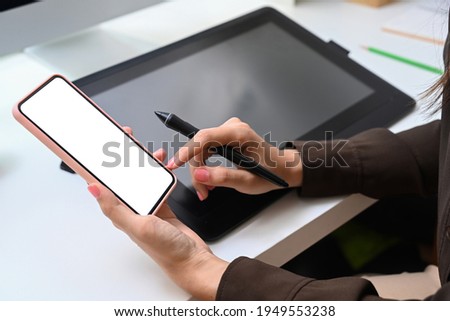 Close up view of female graphic designer holding digitizer pen and using mobile phone at workplace.