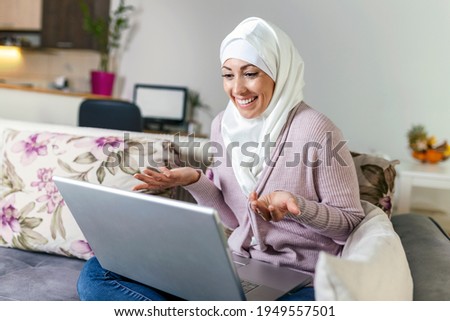 Young Muslim woman having video call via laptop at home. Happy smiling Muslim woman sitting on sofa, couch and using laptop at living room at home, learning language, video calling.