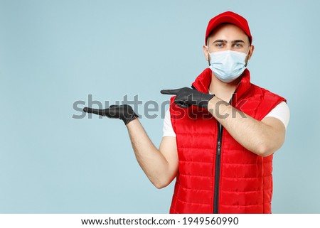 Delivery guy employee promoter man in red cap white t-shirt vest uniform sterile face mask gloves work courier service on lockdown coronavirus flu showing workspace isolated on pastel blue background