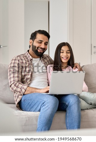 Happy indian family young father with teen child daughter having fun using laptop computer at home watching tv movie, browsing internet, e learning, laughing, sitting on sofa in modern living room. Royalty-Free Stock Photo #1949568670
