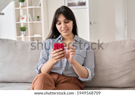 Smiling young indian woman using smart phone sitting on sofa at home. Happy lady holding cell phone doing online shopping, ordering delivery in mobile app, checking social media or dating application.